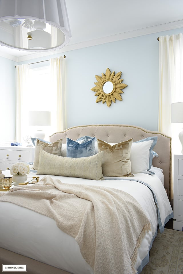 Upholstered bed with brass nailhead trim, layered with white bedding and luxe velvet pillows.