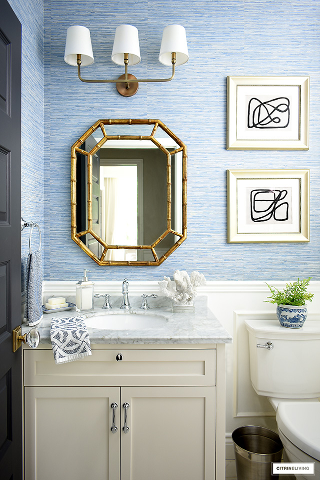 Blue and white coastal-chic bathroom with white vanity, silver hardware and faucet, gold mirror and lighting and blue grasscloth wallpaper.