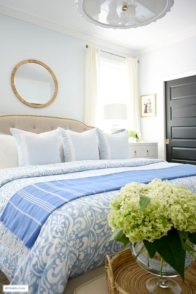 Summer bedroom decorated with pretty light blue and white bedding, rattan mirror and tray, and fresh hydrangeas.