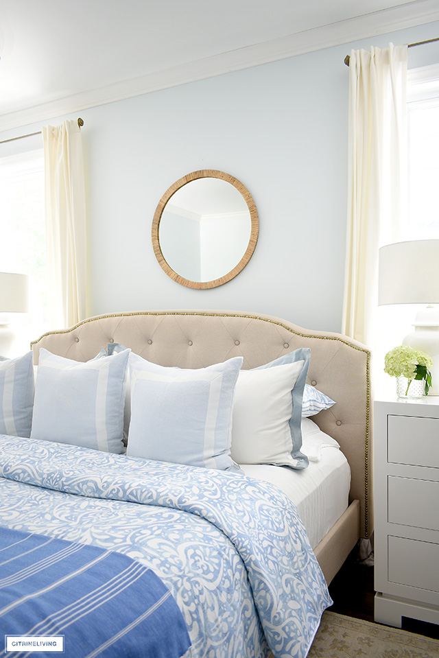 Beautiful bedroom decor with light blue and white bedding, upholstered bed, round rattan mirror.