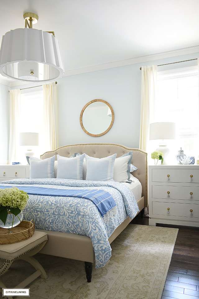 Summer bedroom decor with light blue and white bedding, rattan mirror over the bed and fresh hydrangeas