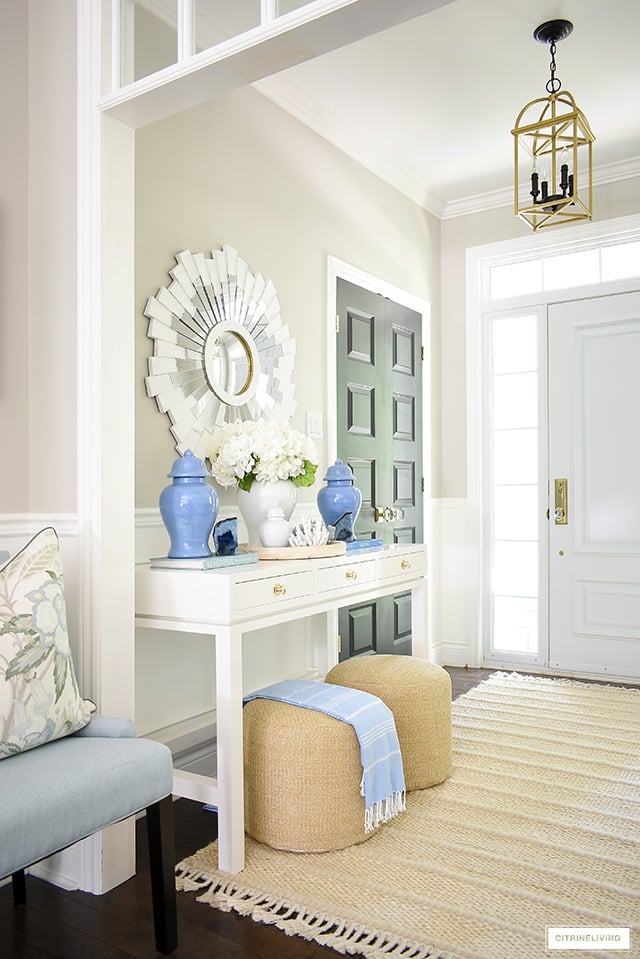 Summer entryway with white console table, blue ginger jars, white sunburst mirror and woven poufs and rug.