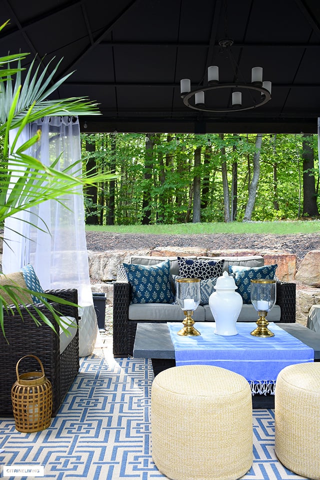 Outdoor gazebo with rattan furniture blue and white pillows and rug, woven poufs and white drapes.