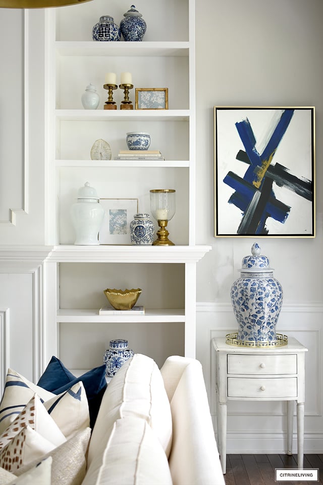 Bookshelff styling ideas with blue and white ginger jars, solid ginger jars, brass hurricane lanterns, and beautiful objects to create interest and tell a story. Modern art accents the color scheme.