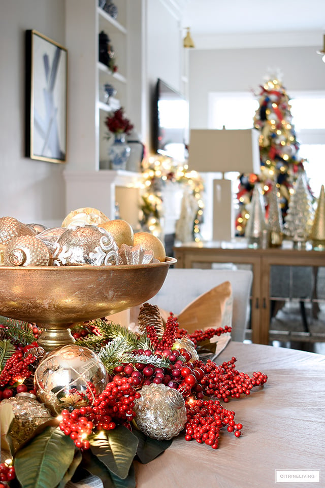 A pretty Christmas centerpiece with ornaments and red berries with magnolia leaves sits on a dining room table, with a view into a living room with garland and Christmas tree.