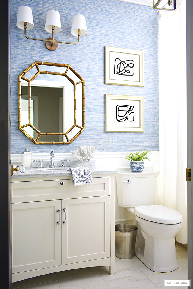 Small bathroom with blue grasscloth wallpaper, white vanity, gold mirror and chrome hardware.