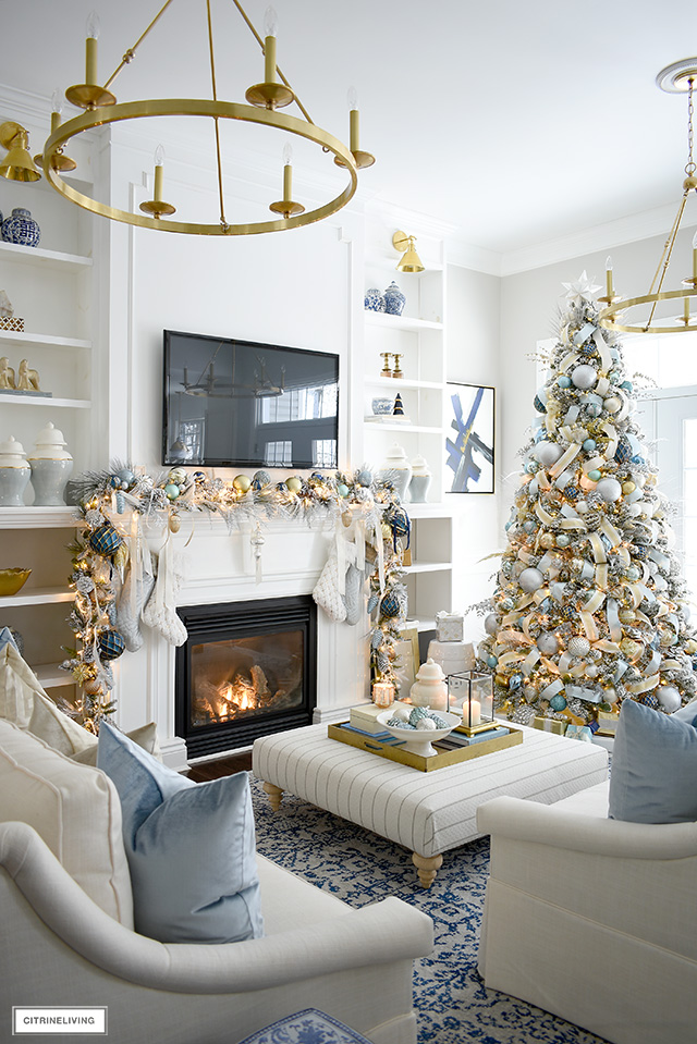 Light blue, gold and white Christmas decorated living room featuring a flocked tree and garland adorned with ornaments and ribbon.