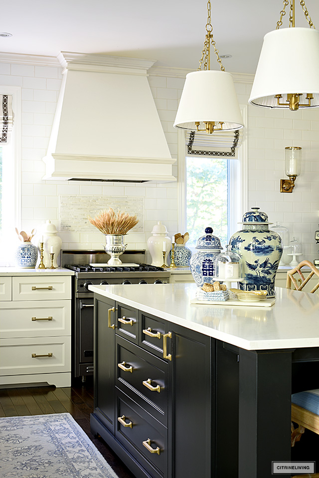 Kitchen styled for fall with a dried wheat arrangement on the stove, and blue and white ginger jars on the island, gold candlesticks are a warm seasonal accent.
