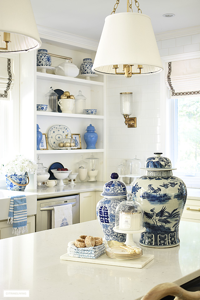 Kitchen island styled with two blue and white ginger jars, and open shelves with a collection of ginger jars, platters and apothecary jars. Gold faux apples are a seasonal accent.