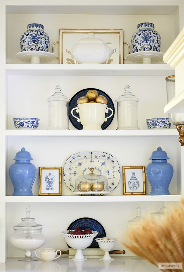 Kitchen shelves decorated for fall with blue and white chinoiserie accents, ginger jars, platters and faux gold apples.