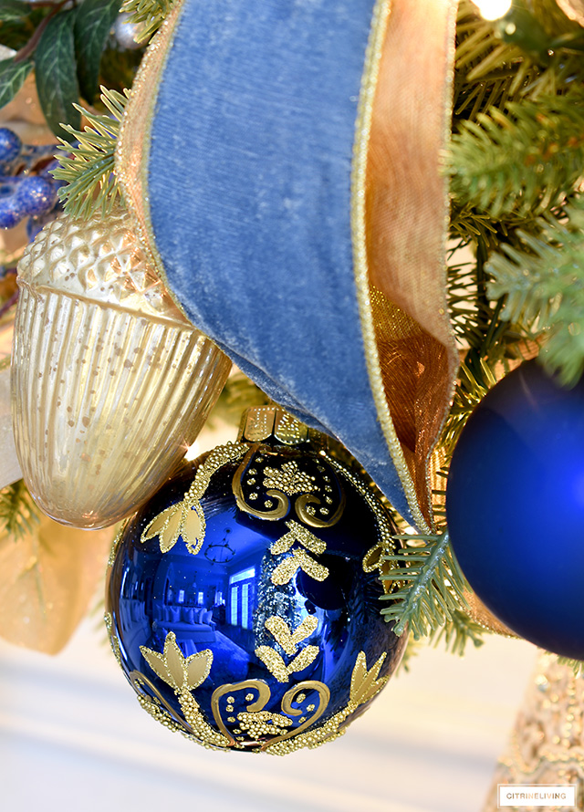 Blue and gold hand-painted Christmas ornament hangs on a green garland with other blue and gold ornaments and ribbon.