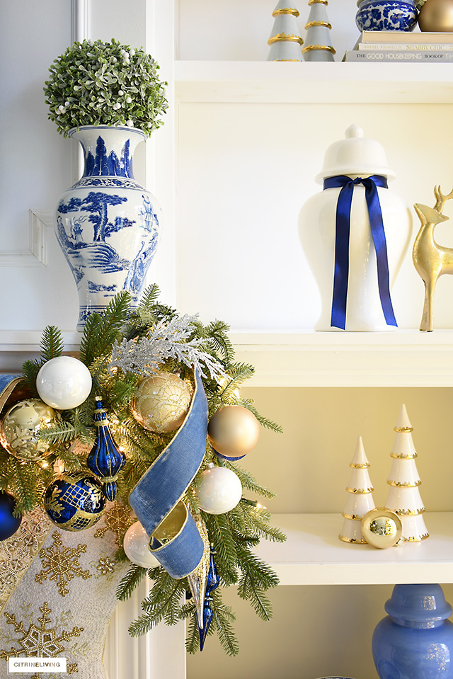 Blue and white chinoiserie vase with green frosted topiary ball sits on a mantel above a gorgeous green garland decorated with blue and gold.