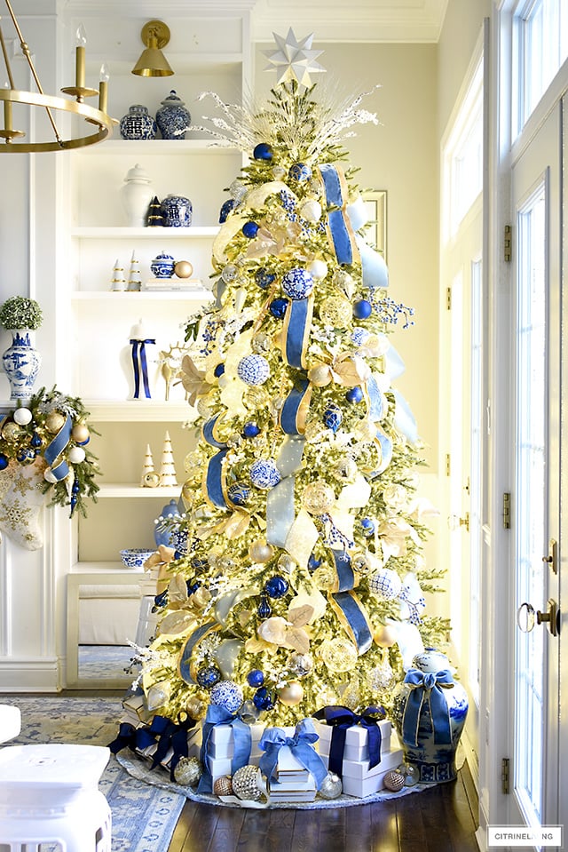 Stunning pre-lit gold and blue decorated Christmas tree standing in front of simply decorated bookshelves for Christmas.