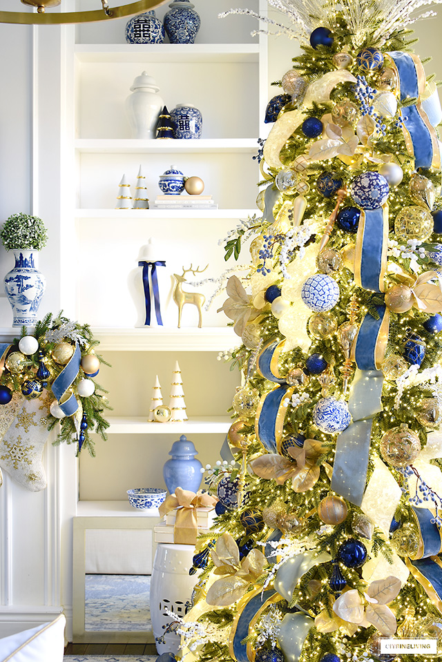 Living room shelves decorated for Christmas with blue, white and gold accents, anelegantly decorated gold and blue Christmas tree stands to the right.