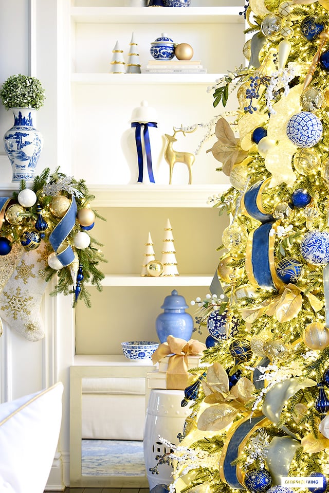 A gorgeous glimpse of a Christmas decorated living room - bookshelves with blue and gold Christmas decor, a faux green garland and Christmas tree adorned with blue and gold ribbon, picks and ornaments.