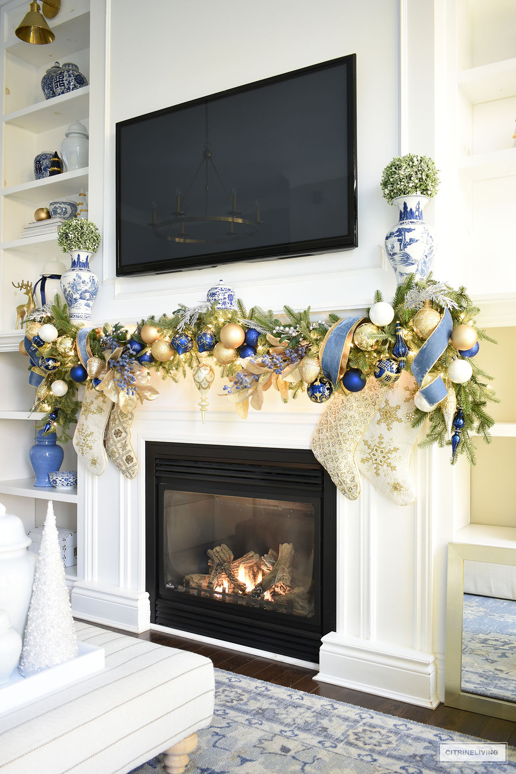 Fireplace decorated with a swagged garland adorned with blue and gold decorations and ribbon for christmas. Blue and white chinoiserie vases with topiaries balls sit above on each end flanking the tv.