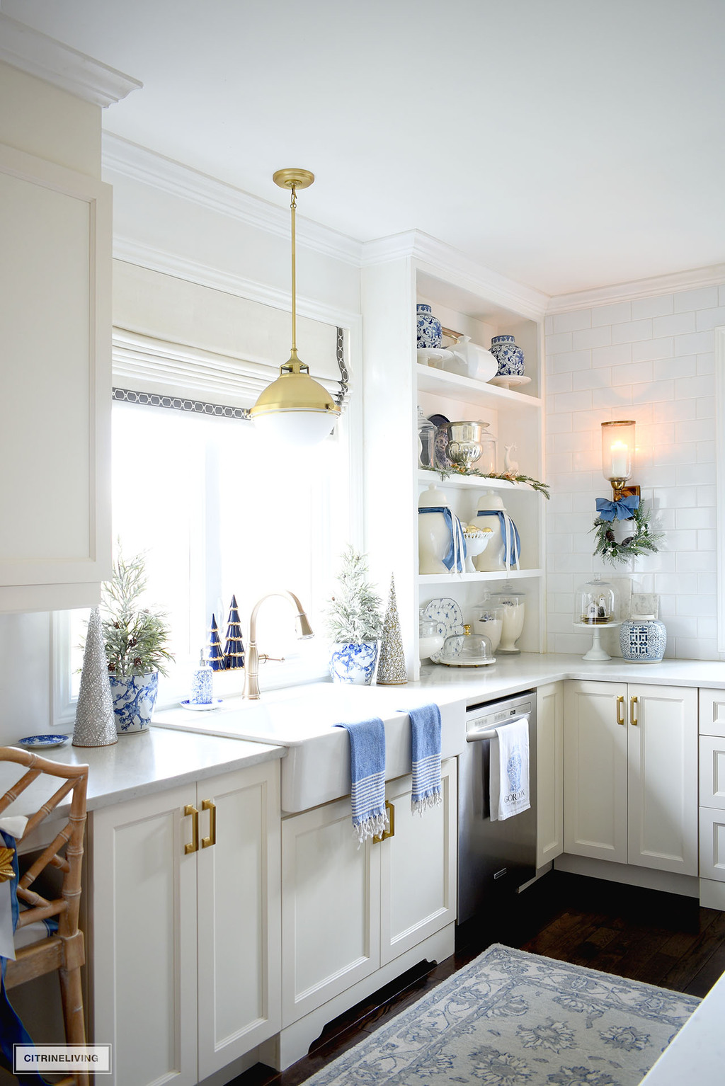 White kitchen farm sinks decorated for Christmas with blue and white accents and trees, open shelves are styled with a mix of white and gloss jars and blue and white accents.