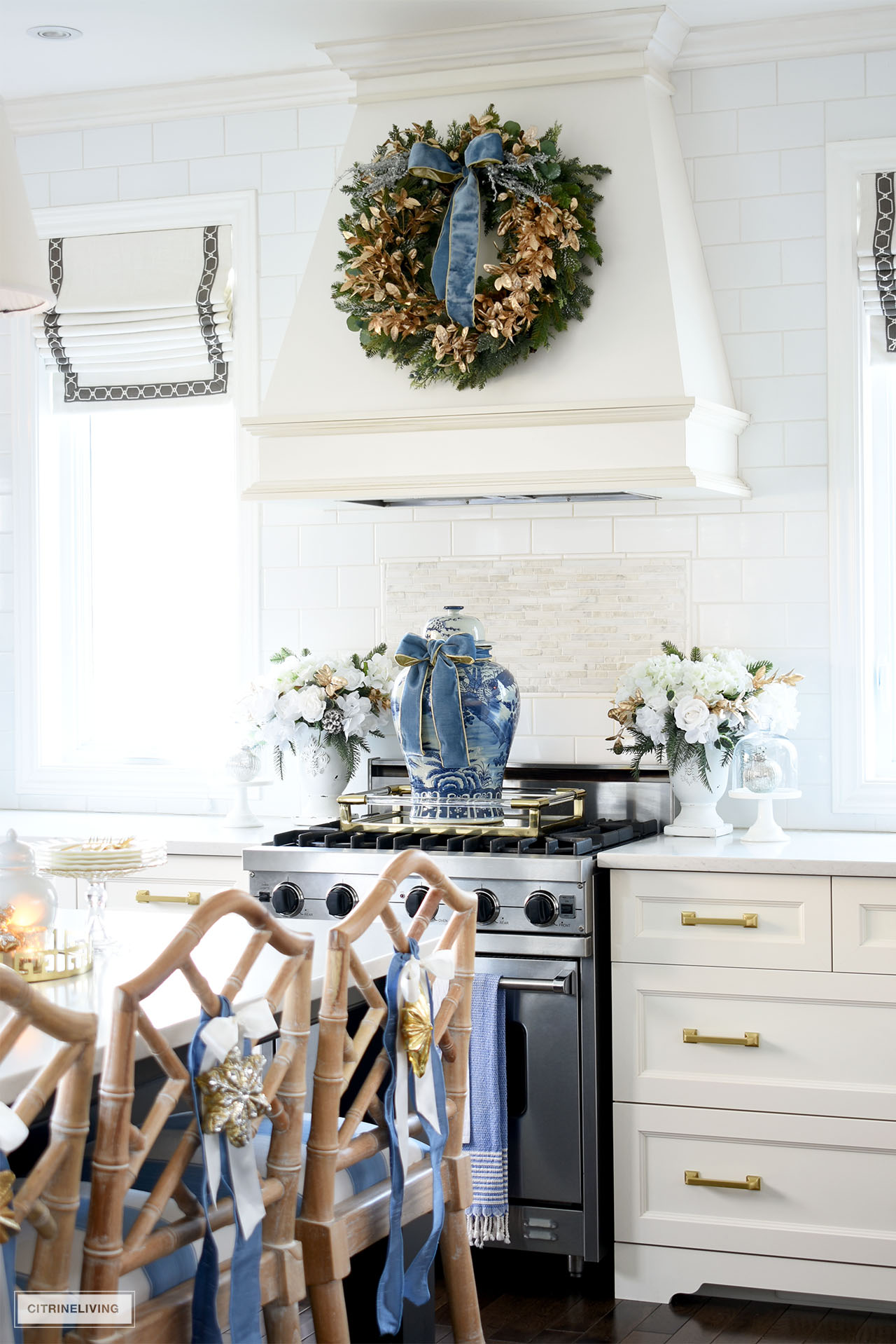 Range hood styled with a lush green and gold wreath with blue velvet ribbon hangs above an oversized blue and white ginger jar and gold tray on the stove. Gorgeous white, green and gold floral arrangements sit on either side of the stove.