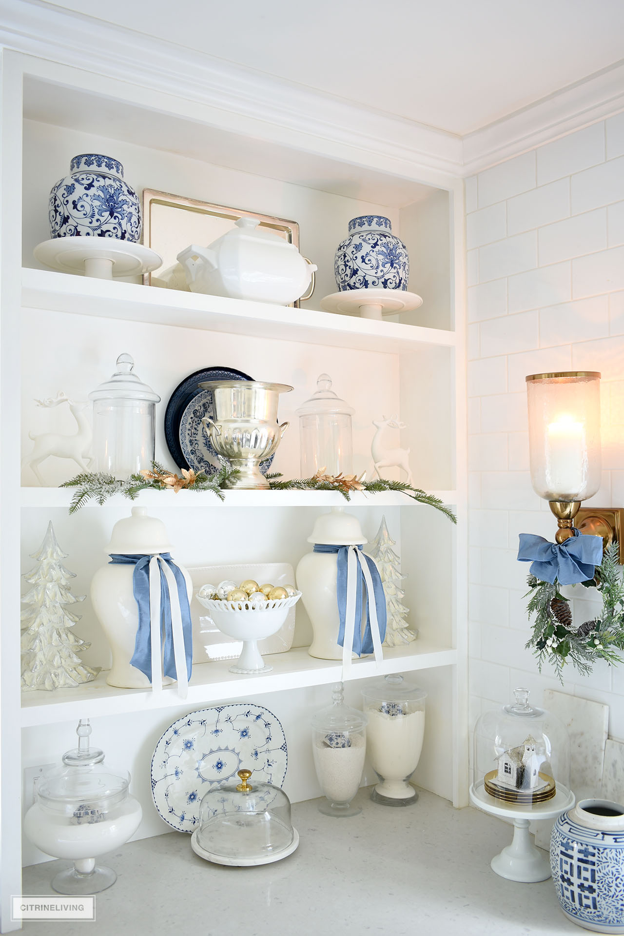 Open builtin kitchen shelves styled for Christmas with blue, white, greenery and gold touches.