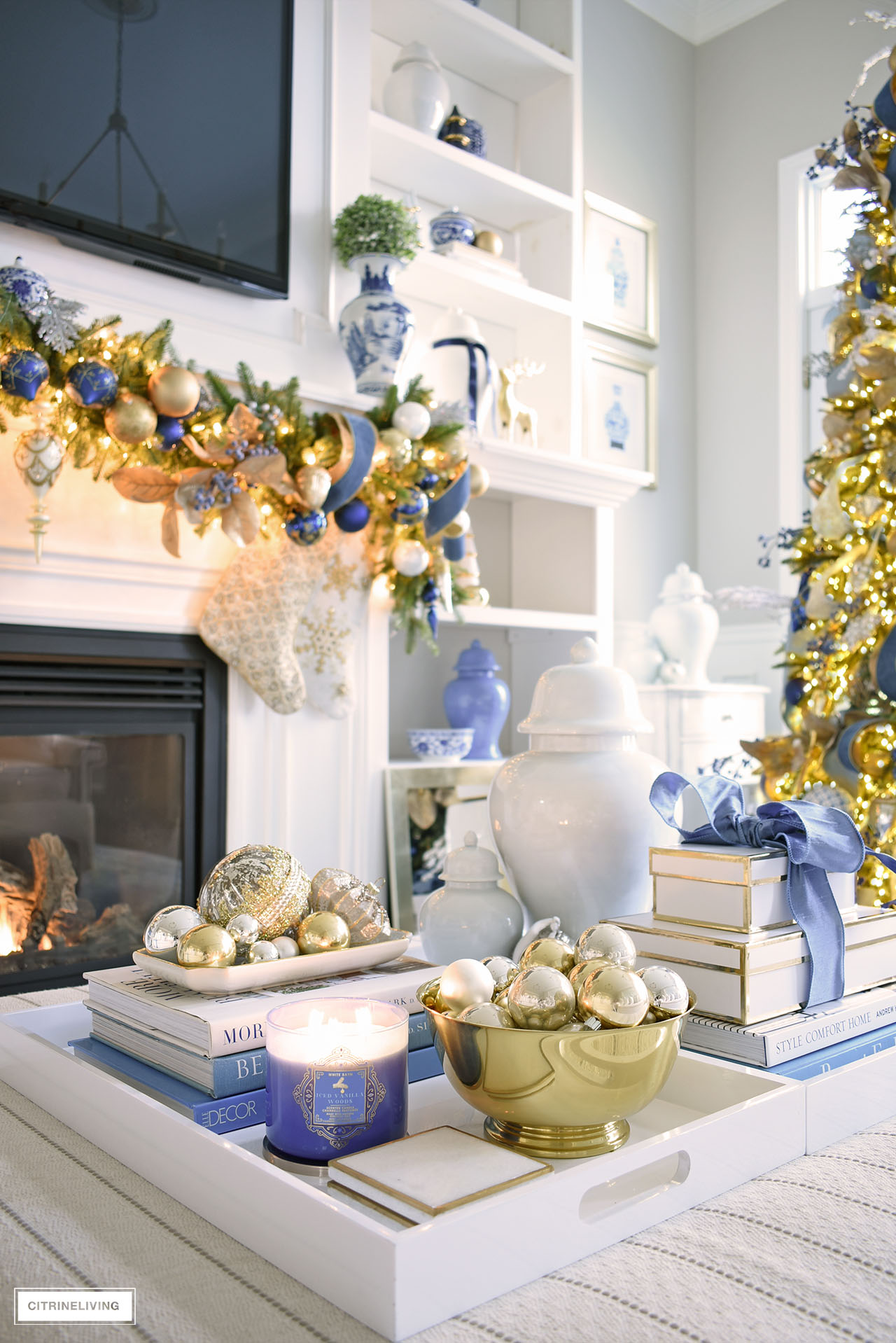 Living room ottoman styled for Christmas with white trays and blue and gold accessories, ornaments, ginger jars, candle and gift box.