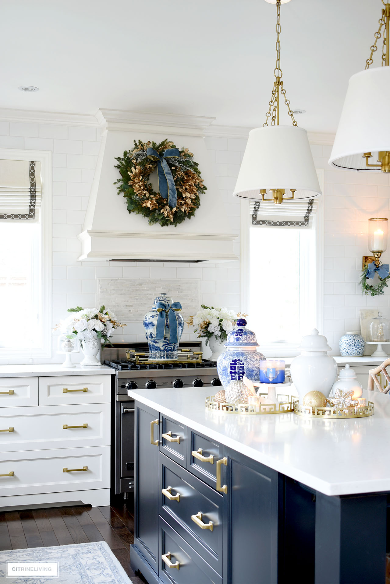 Kitchen island styled for Christmas with ginger jars, candles, ornaments and trays. A large ginger jar on the stove with a blue velvet ribbon is an elegant detail along with a green and gold wreath hung above on the hood.