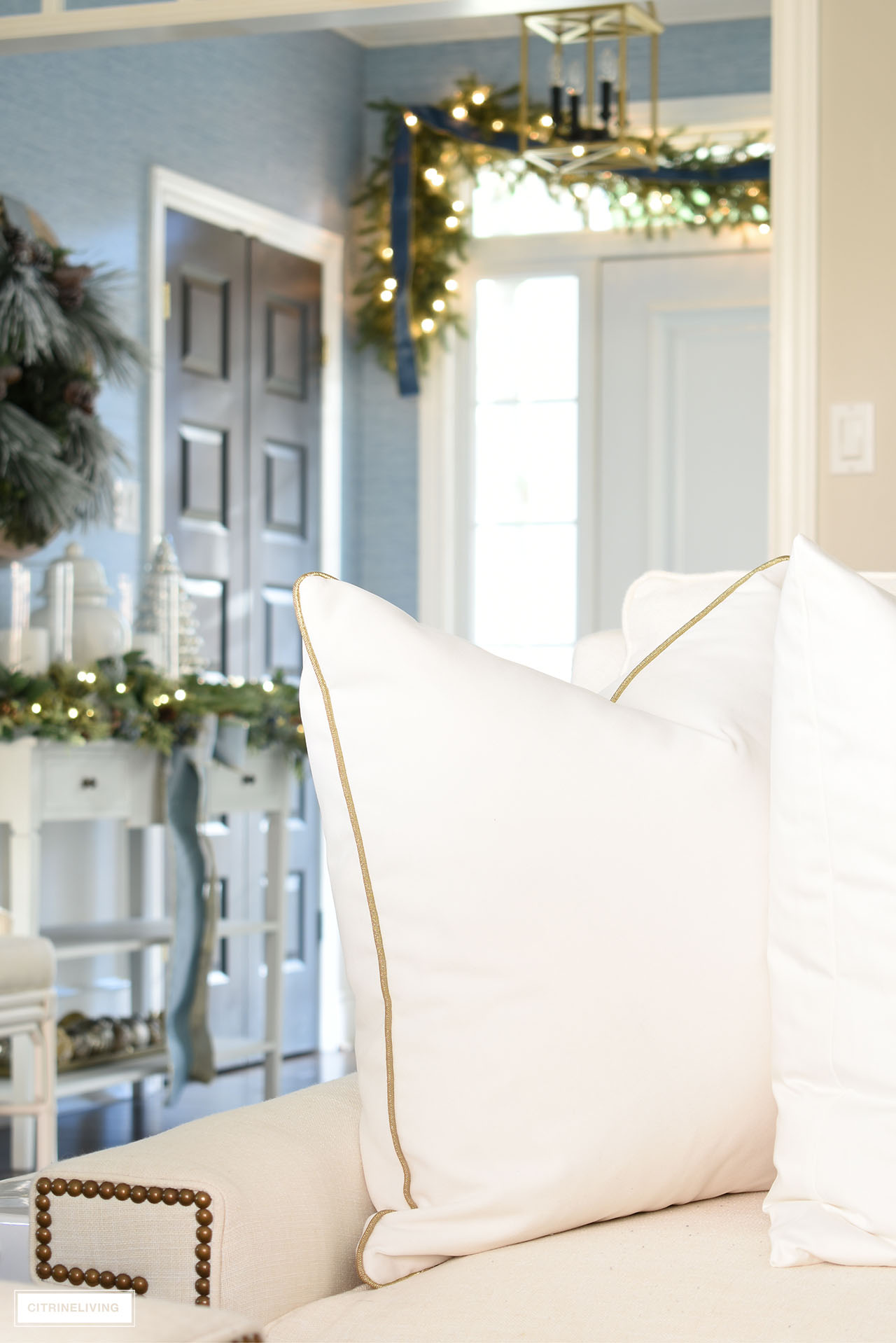 White velvet pillow with gold piping detail on a white sofa. Christmas garland hangs in the entryway in the background.