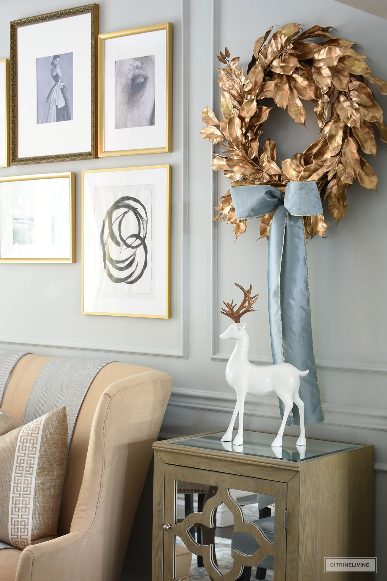Good leaf wreath with a silver-blue taffeta bow hangs on a gallery wall with a simple and elegant white and gold reindeer standing below on top of a mirrored cabinet.