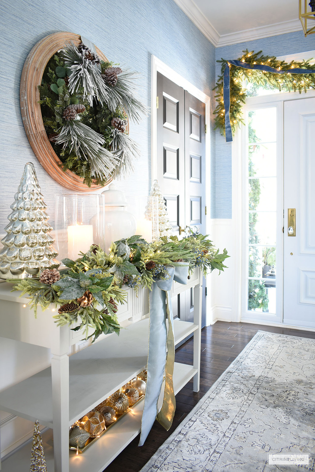 Entryway decorated for Christmas with green wreath, garlands, blue ribbons and silver and gold accessories.