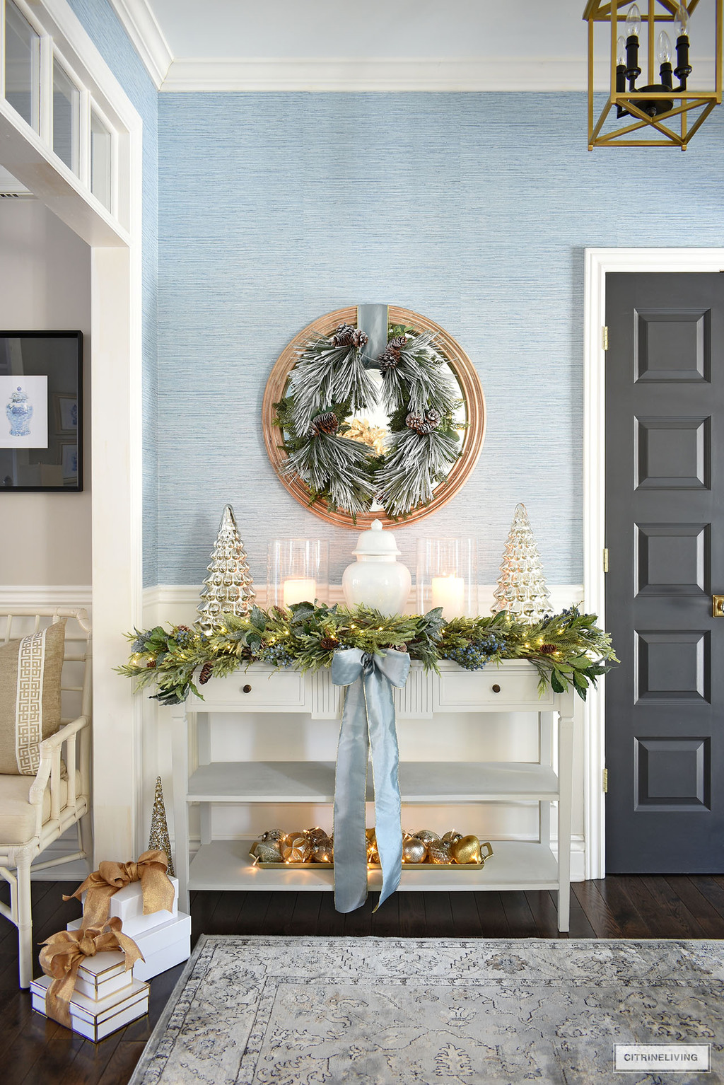 Elegant Christmas entryway decor with lush greenery, blue ribbons, frosted green wreath, ginger jar, hurricanes and mercury glass trees.