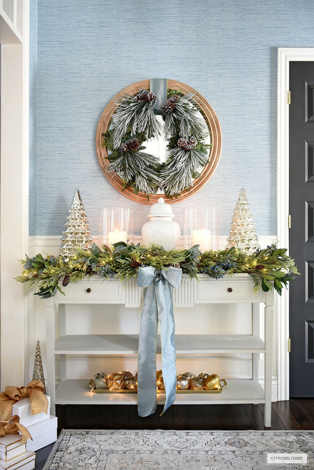 Console table styled for Christmas with a light blue ginger jar, glass hurricanes and mercury glass trees on either side. Lush greenery garland and frosted wreath complete the look. 