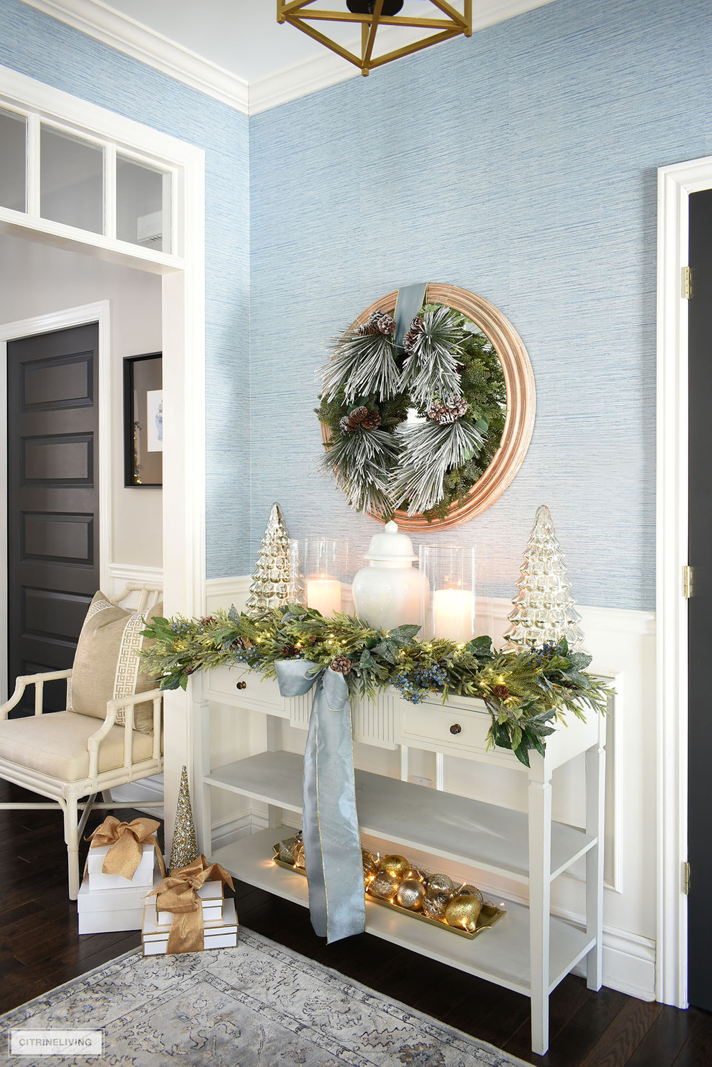 Console table decorated for Christmas with greenery, blue ribbon, ornaments, light blue ginger jar and mercury glass trees.