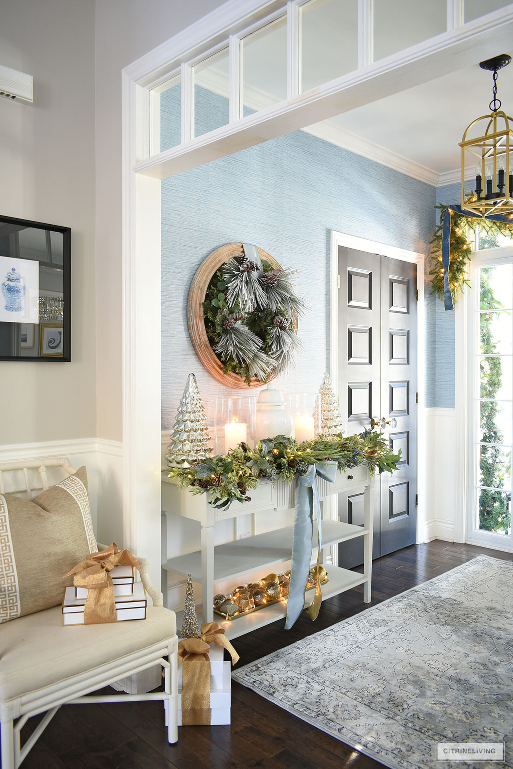 Christmas entryway decor with green garland and wreath, mercury glass trees, hurricane lamps and gold and silver ornaments.