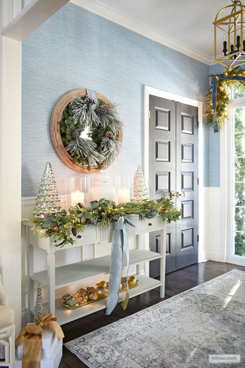 Entryway decorated for Christmas with greenery, blue ribbon, mercury glass trees, silver and gold ornaments.