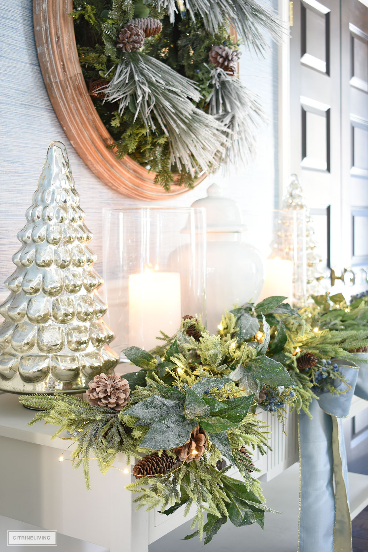 Close up of Christmas console table with greenery garland, mercury glass trees, hurricanes and ginger jar.