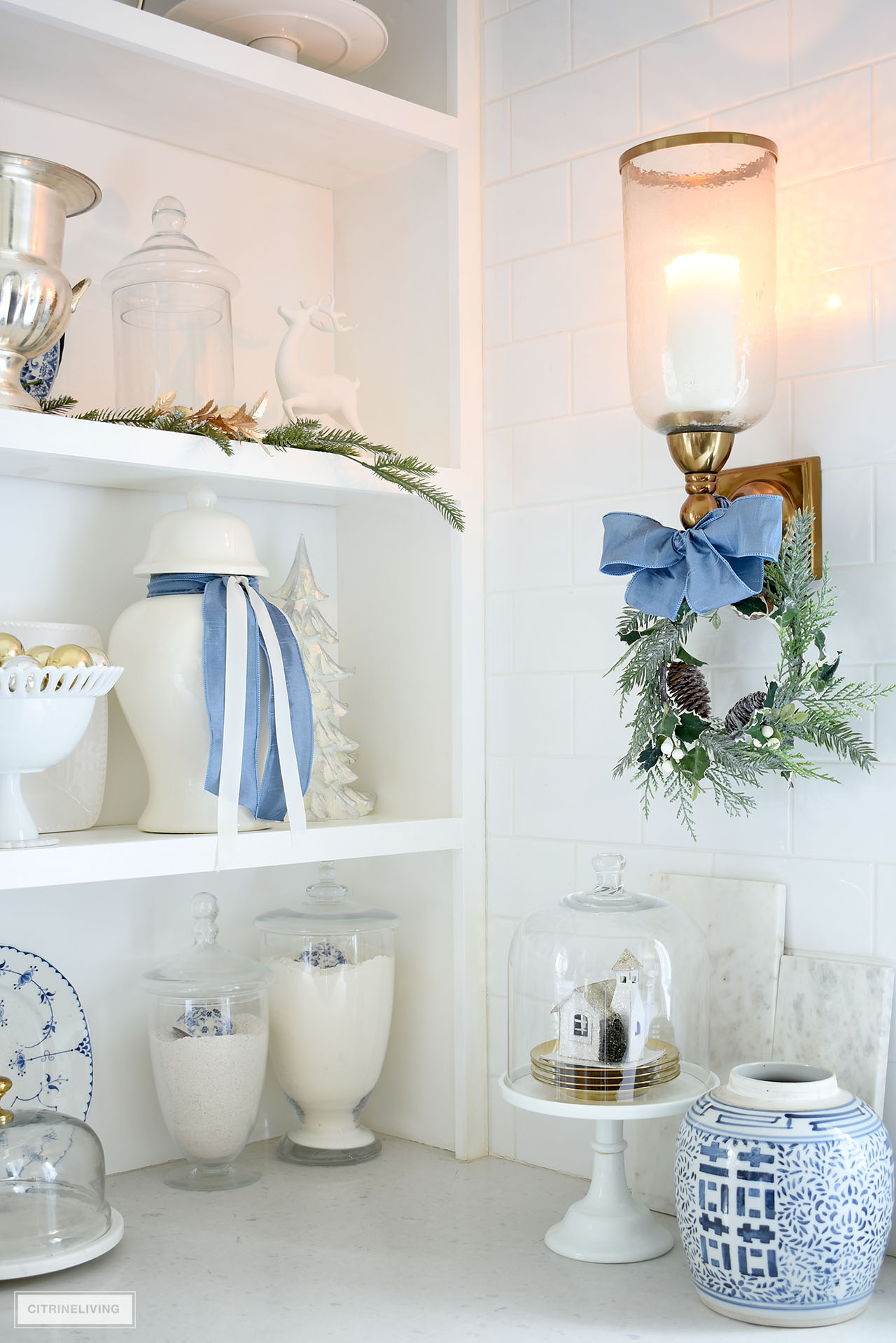 Corner detail of open kitchen shelves and gold wall sconce, decorated for Christmas with blue and white accents and greenery.