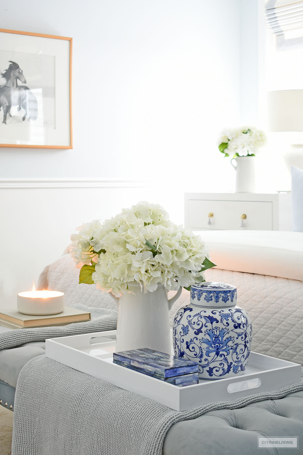 A chic white tray styled with a white vase filled with faux hydrangeas, a blue and white ginger jar and blue decorated box sit at the foot of the bed.