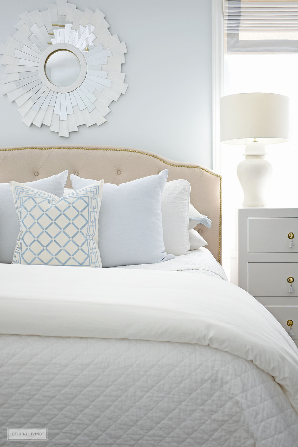 A beautiful layered bed with white quilt, soft blue and white throw pillows, a starburst mirror above the bed and a pretty ginger jar lamp on the bedside dresser.
