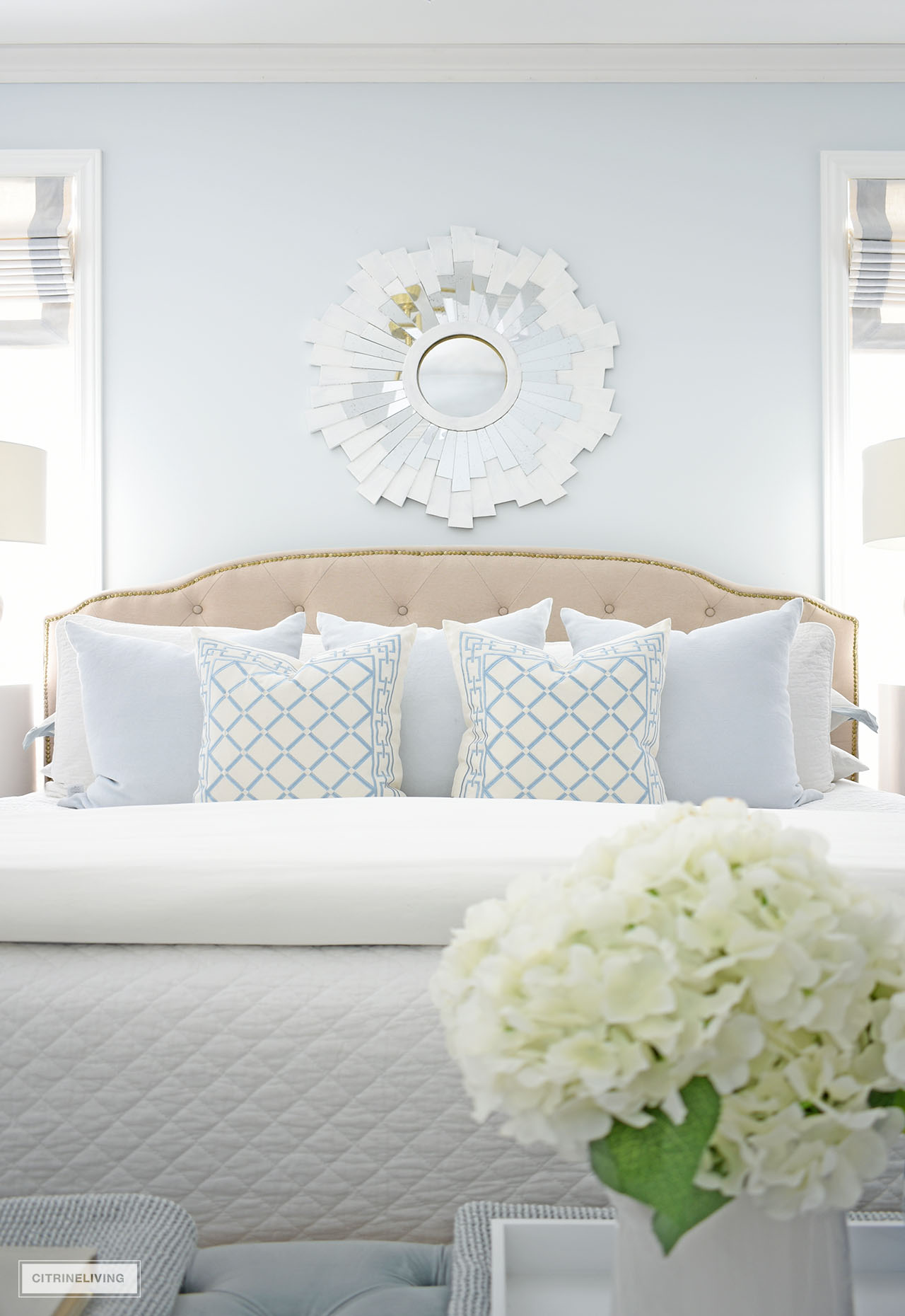 A gorgeous layered bed with light blue and quilted white bedding, and a chic white starburst mirror above the bed.