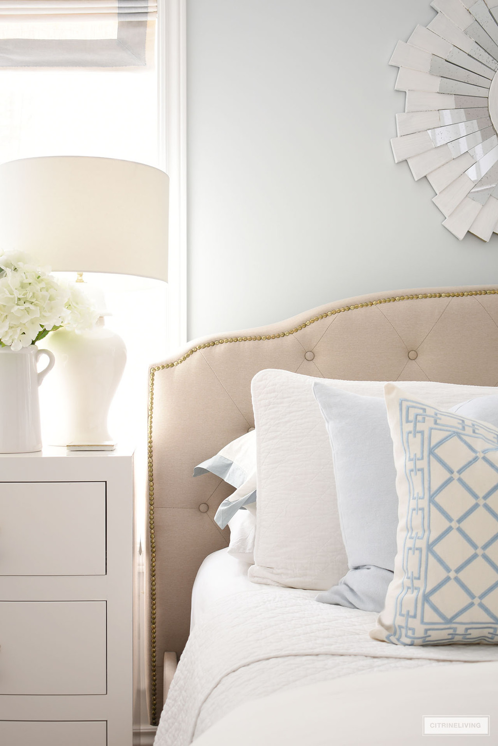 Beautiful layers of white and light blue bedding and pillows on an elegant upholstered bed with nailhead trim.