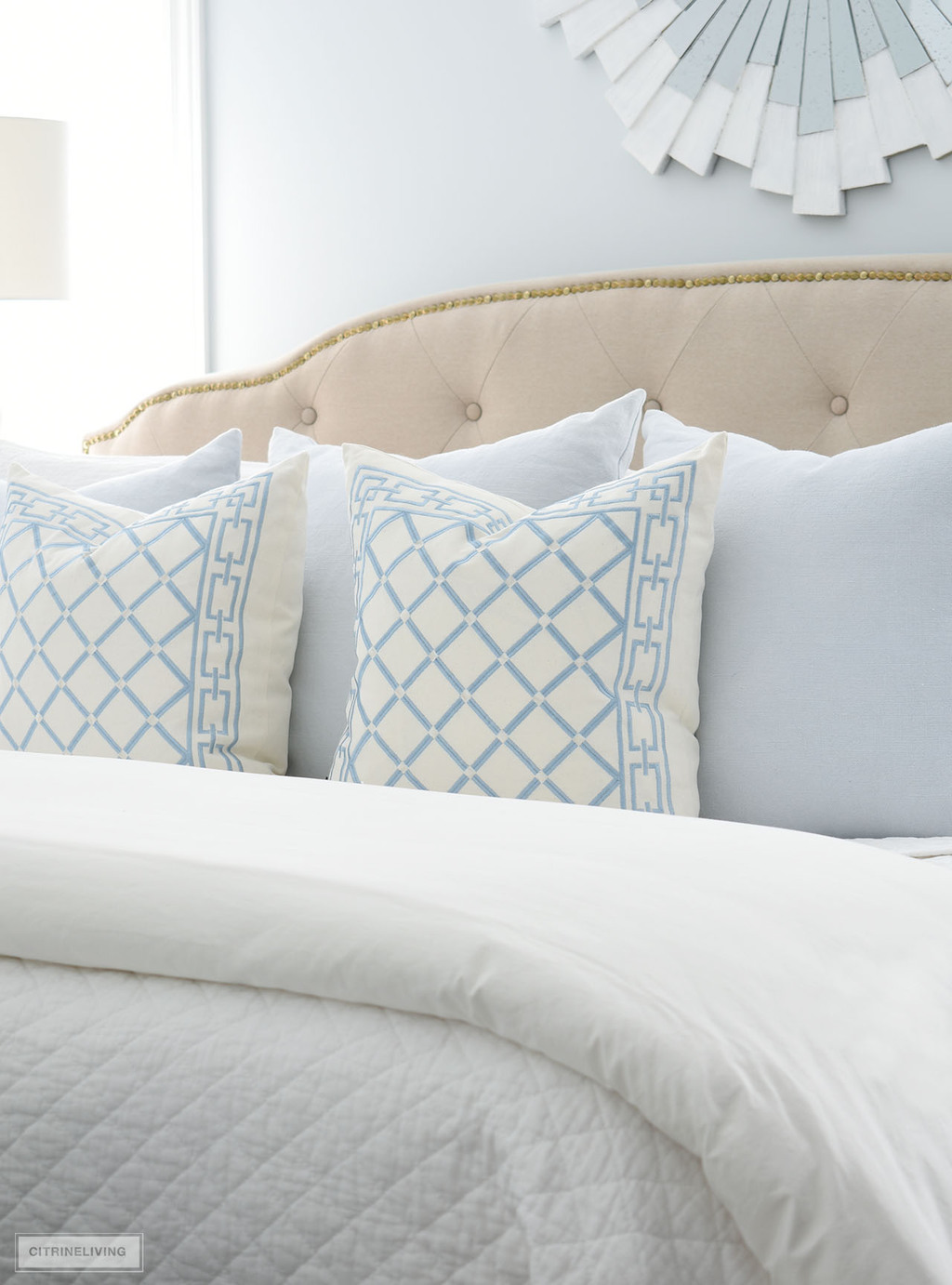 Light blue and white embroidered pillows in a diamond pattern, layered with pretty pale blue pillows styled on an upholstered bed.