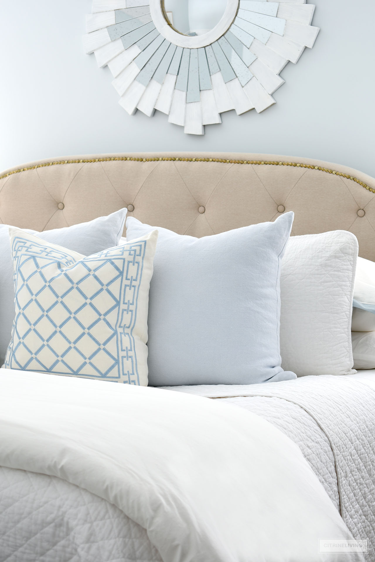 Layers of light blue and white embroidered pillows in a diamond pattern, layered with pretty pale blue linen pillows styled on an upholstered bed.