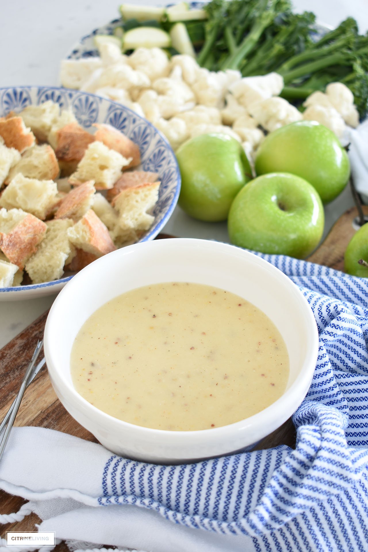 Cheese fondue in a white bowl served with crusty homemade bread, green apples, cauliflower and broccolini.