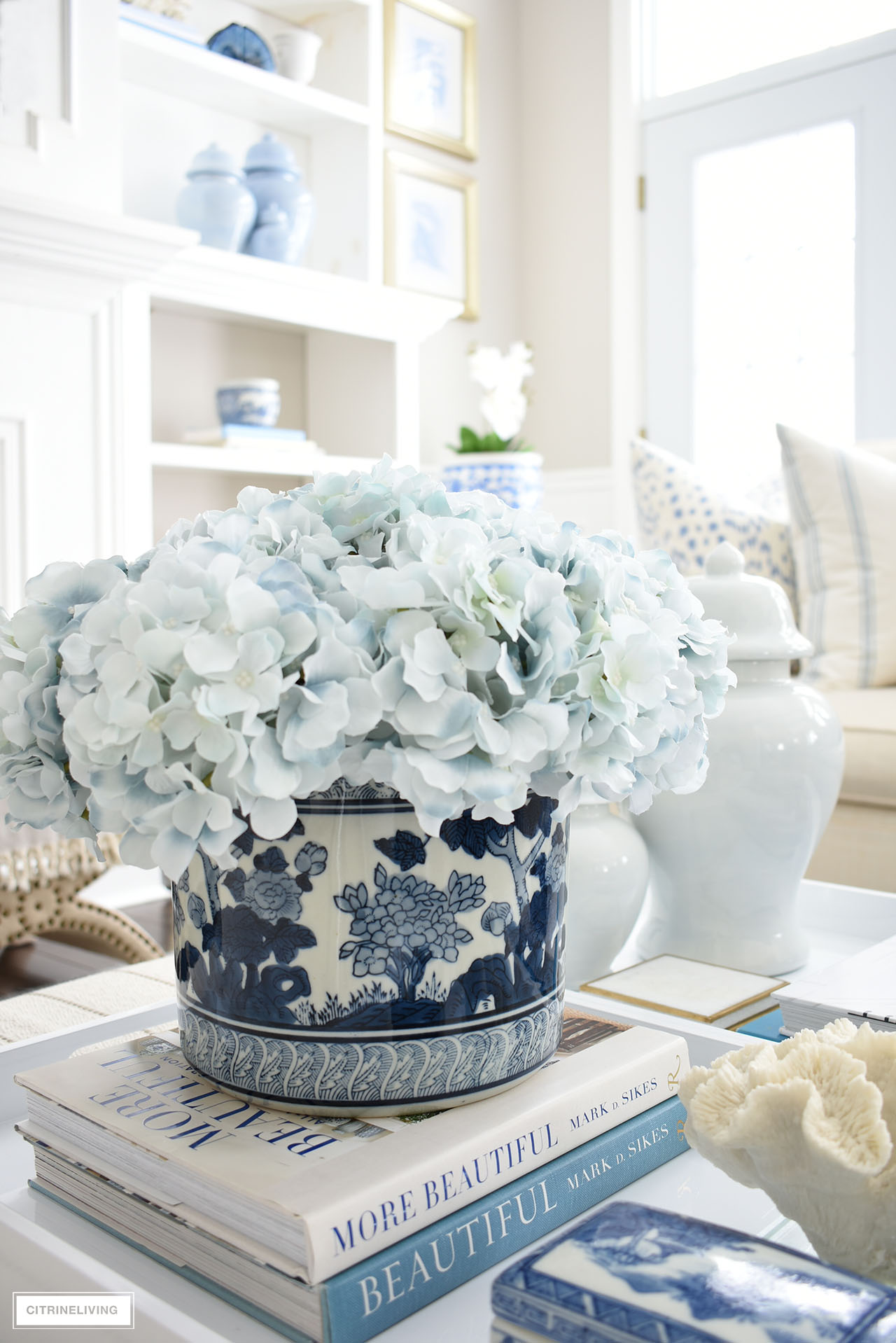Spring coffee table decor with light blue hydrangeas arranged in a blue and white planter, styled atop decorating books on a white tray.