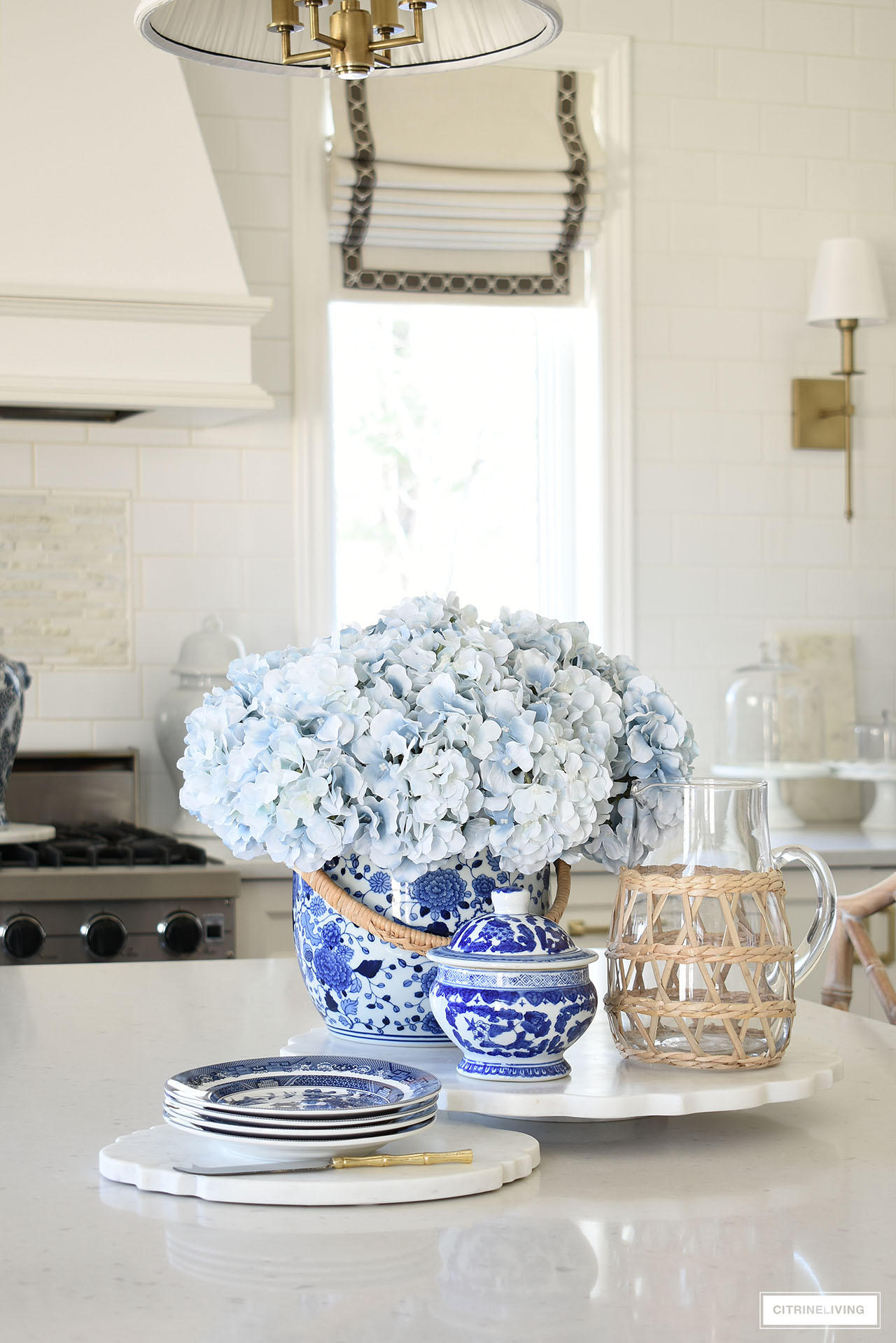 A pretty kitchen island vignette with blue and white chinoiserie, light blue hydrangeas, marble trays and a woven wrapped glass pitcher.