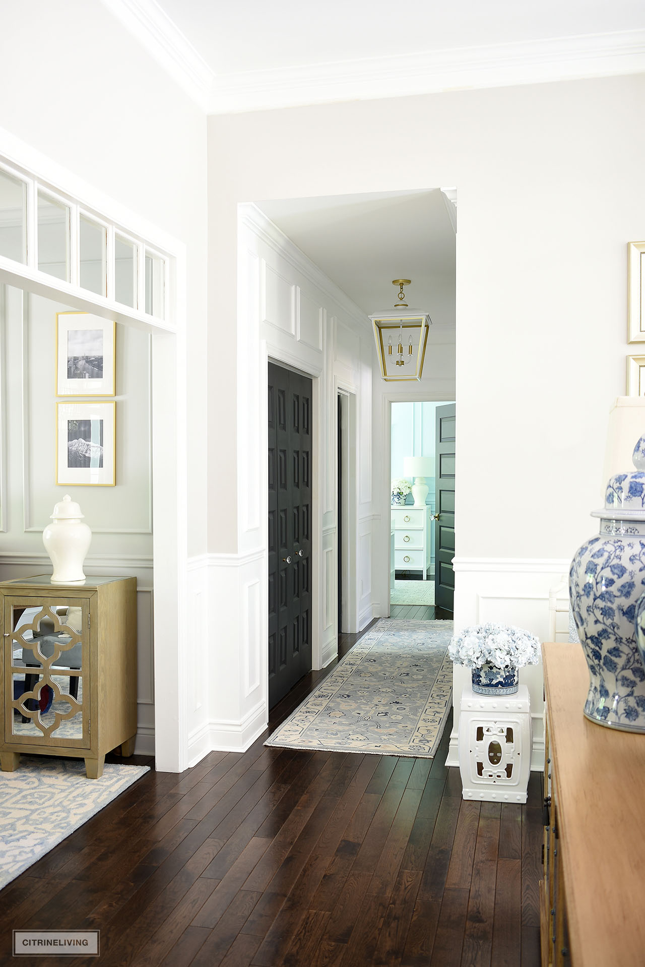 Open concept home with a view down a white painted hallway with moldings, large white and gold pendant light, and light blue oushak rug