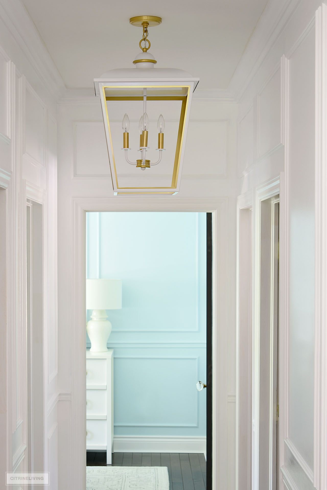 An oversized lantern pendant light in white and gold hangs in a small hallway painted Ultra Pure White by Behr.