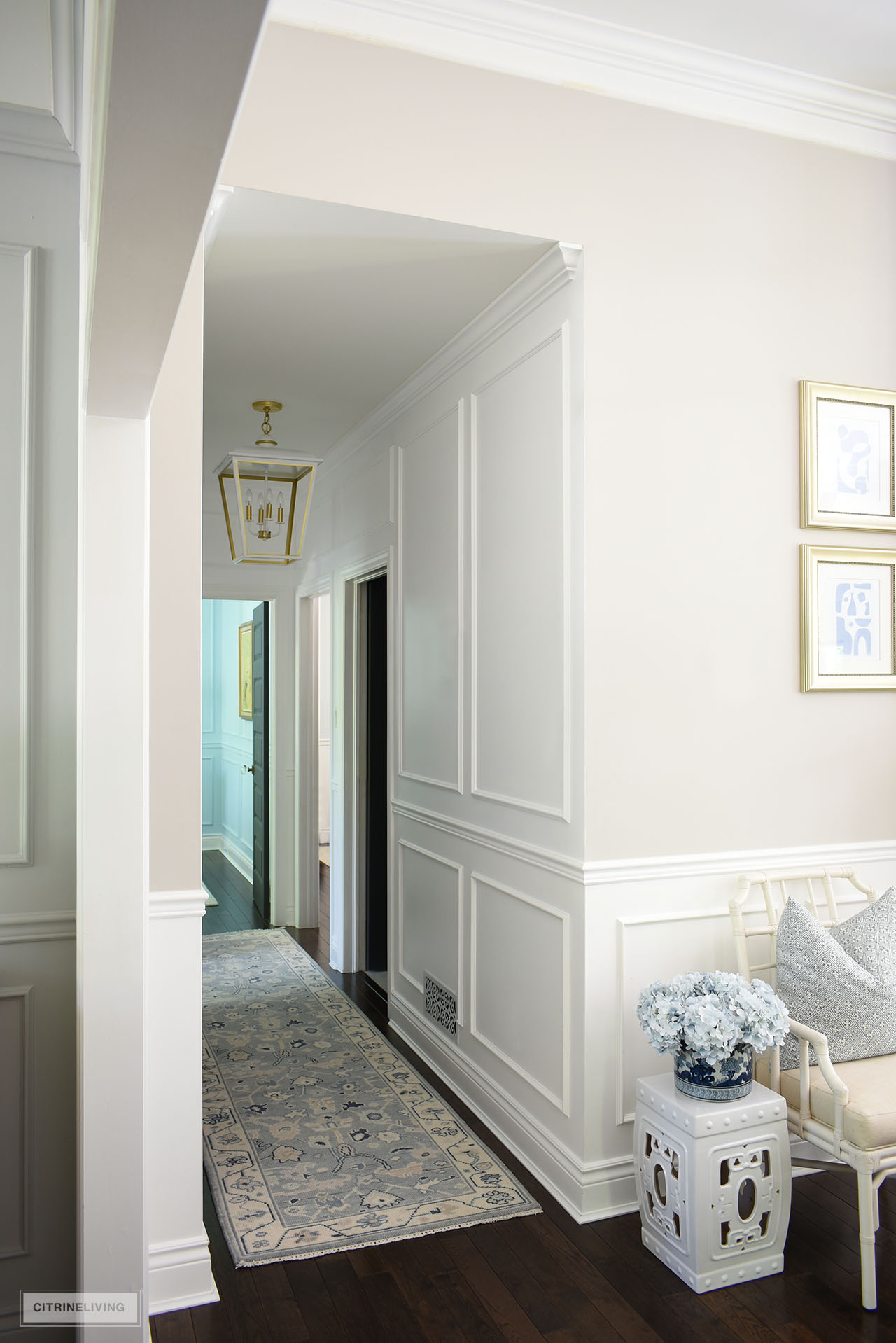 White painted hallway featuring wall moldings, light blue oushak rug ad large white ad gold pendant light.