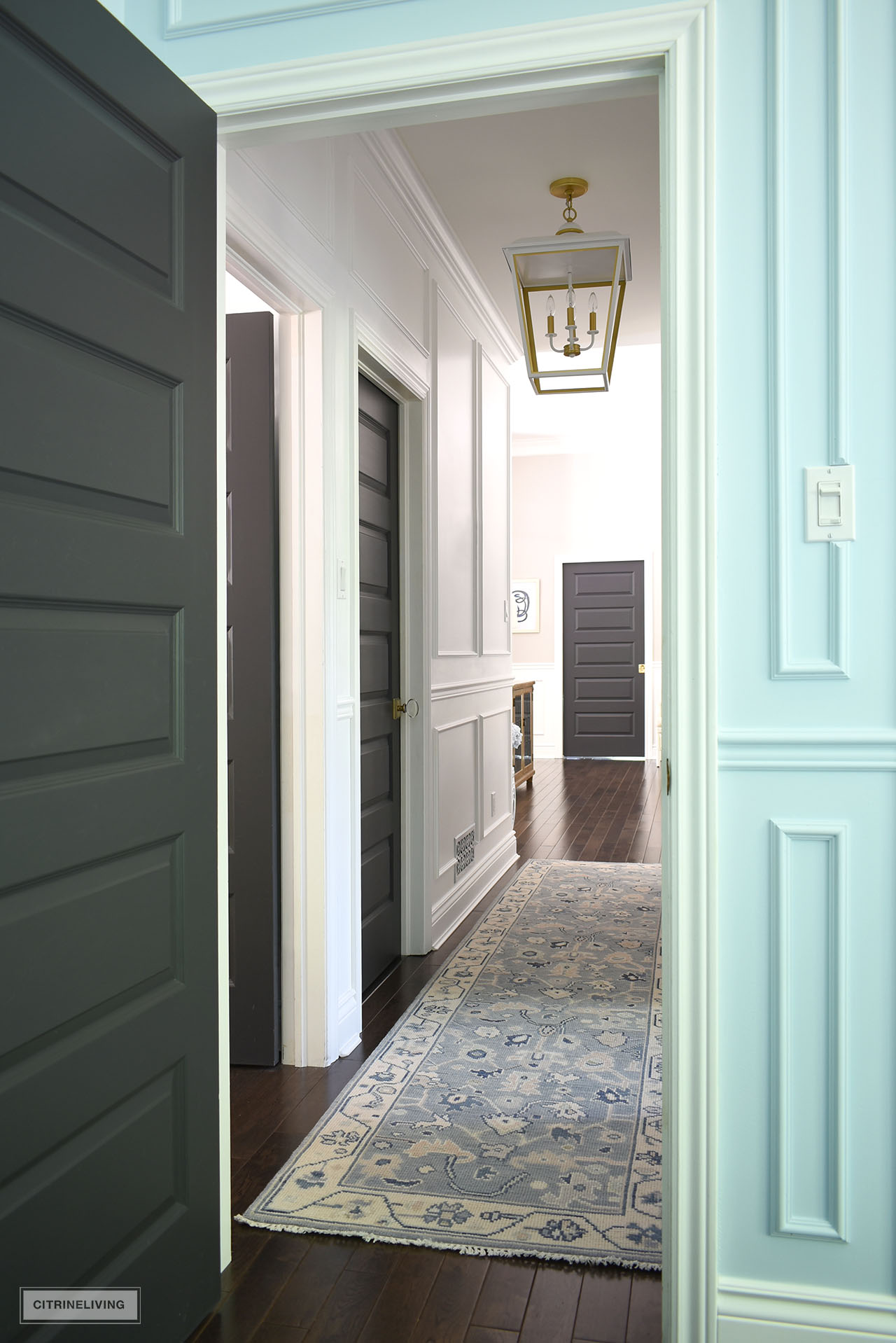 A view down a small but elegant hallway painted white, with wall moldings, crown moldings, a large oversized pendant light and light blue oushak runner.