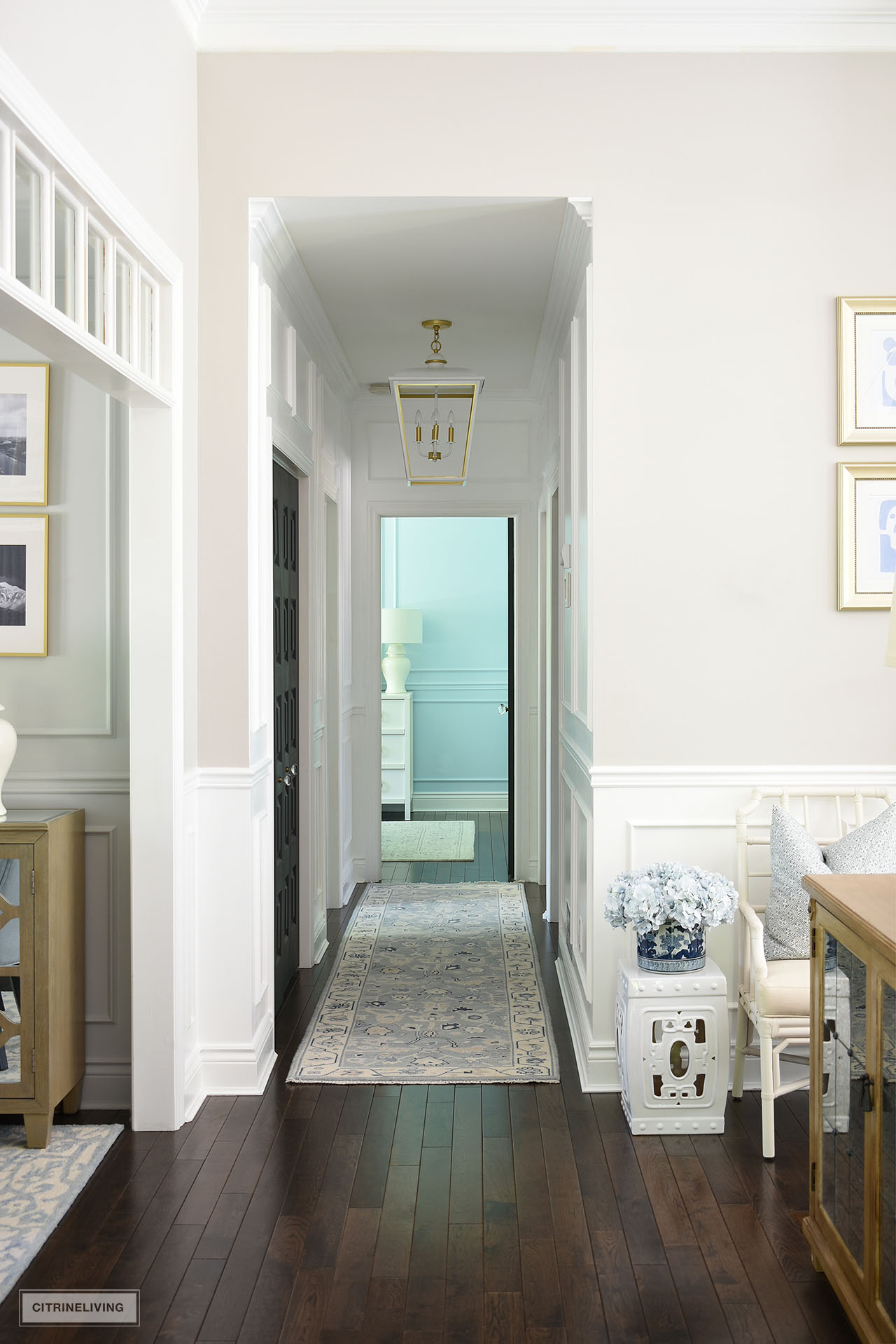 A view of a narrow hallway painted white in Behr Ultra Pure White, semigloss paint. Crown and applied wall moldings are elegant and the large oversized pendant light in white and gold is a sophisticated touch. A light blue oushak runner brightens up the dark hardwood floor.