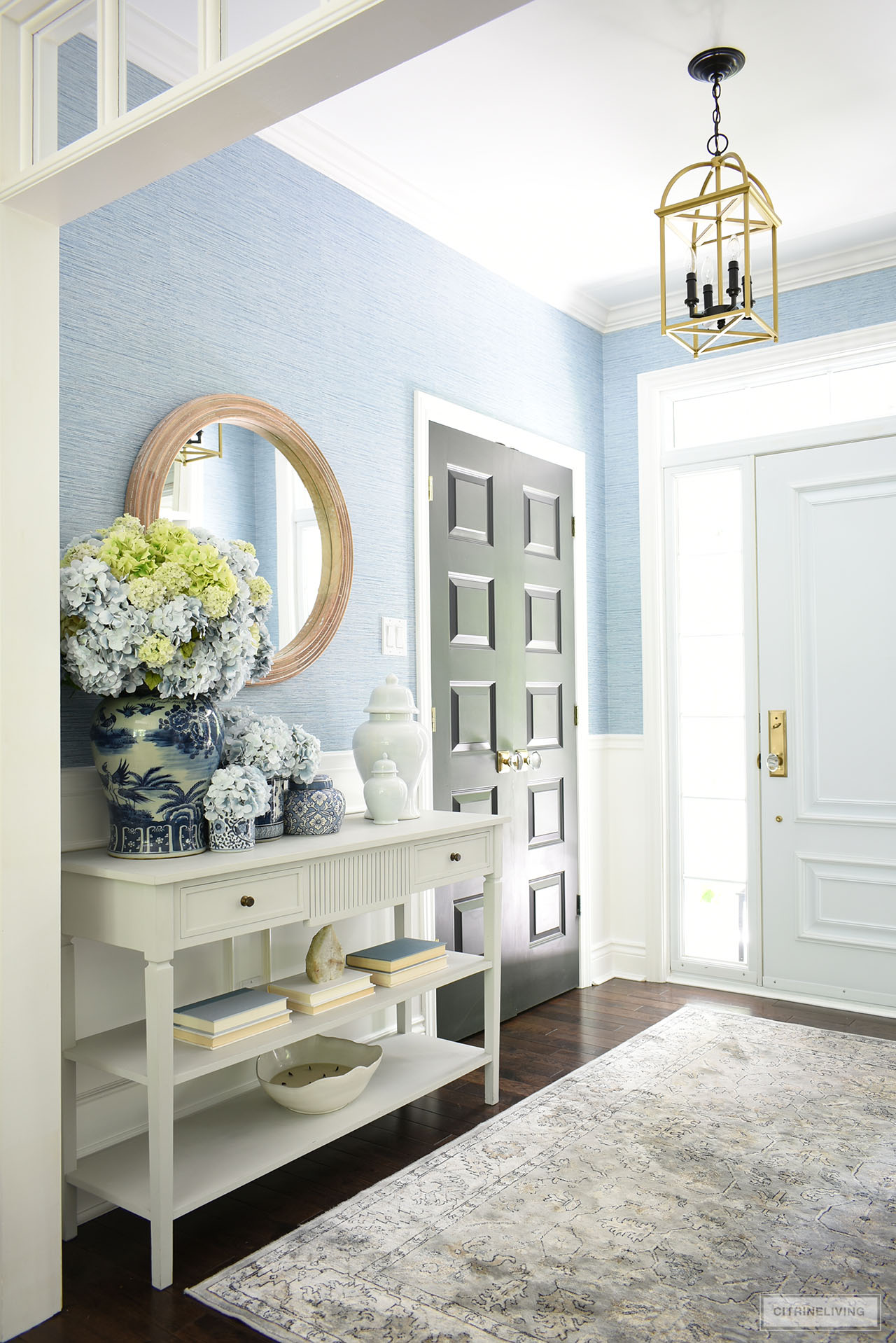 A bright and airy entryway with blue grasscloth is decorated for summer with gorgeous blue and white jars and vases arranged with gorgeous light blue and green hydrangeas.